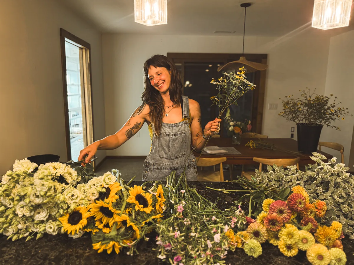 Abby smiling as she arranges bouquets at the counter, a stem of rudbeckia triloba in hand and piles of sunflowers, lisianthus, and zinnias in front of her.