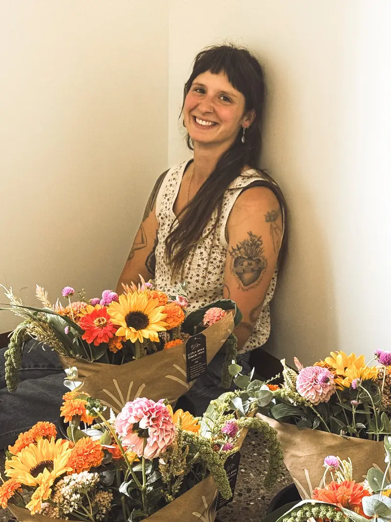 Abby sitting on the floor and smiling, surrounded by finished Little Bend bouquets of sunflowers, zinnias, and dahlias wrapped in kraft paper.