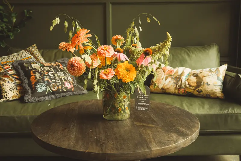 A Little Bend bouquet of dahlias, zinnias, coneflower, and oats in a glass vase on a wooden table in front of a green sofa.